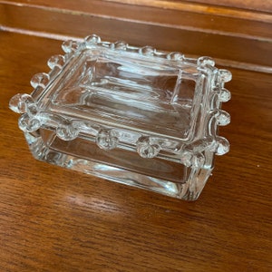 May include: A clear glass trinket box with a square shape and a decorative, beaded edge.