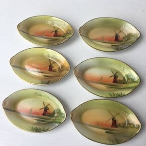 May include: Set of six oval-shaped, cream-colored ceramic dishes with gold trim. Each dish features a hand-painted scene of a windmill with a red roof and a sailboat on a body of water.