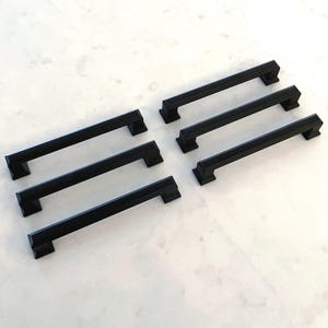 May include: Six black cabinet pulls with a rectangular design are arranged on a white surface. The pulls have a matte finish and a simple, modern aesthetic. They are likely used for drawers or cabinet doors.