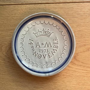 May include: A round, off-white ceramic plate with a blue rim and a scalloped edge. The plate features a crown and the inscription "14th November 1973 A*M". The plate is likely a commemorative item or a decorative piece.