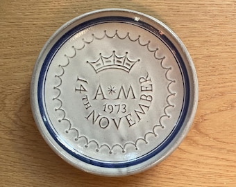 May include: A round, off-white ceramic plate with a blue rim and a scalloped edge. The plate features a crown and the inscription "14th November 1973 A*M". The plate is likely a commemorative item or a decorative piece.