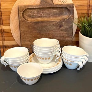 May include: A collection of white ceramic teacups and saucers with a gold floral pattern. The teacups have a curved handle and are stacked in groups. A wooden cutting board and a potted plant are in the background.