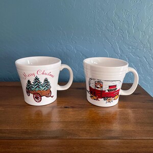 May include: Two white ceramic mugs with Christmas-themed designs. One mug features a cart carrying Christmas trees with the text "Merry Christmas." The other mug displays a red and white camper with pumpkins.