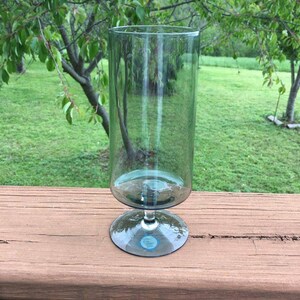 May include: A clear glass vase with a cylindrical top and a round base. The vase is supported by a stem. The vase is sitting on a wooden surface, with a green background of trees and grass.