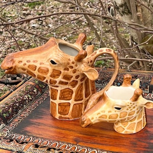 May include: Two ceramic giraffe-shaped pitchers. The larger pitcher has a handle and a long neck, while the smaller one is a creamer. Both are painted in shades of brown and tan, with a spotted pattern.