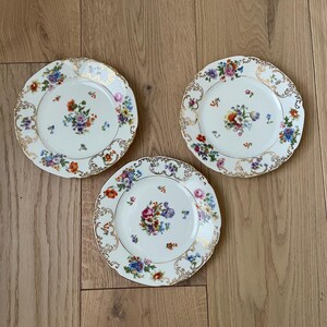 May include: Three white porcelain plates with a floral design. Each plate features a central bouquet of colorful flowers and a gold-colored, ornate border. The plates are arranged on a light brown wooden surface.