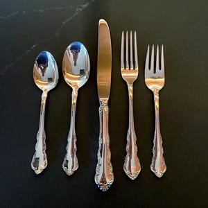 May include: A set of five silver-colored flatware pieces, including two spoons, a knife, and two forks, arranged on a dark surface. The utensils have ornate, detailed handles and reflect the surrounding environment.