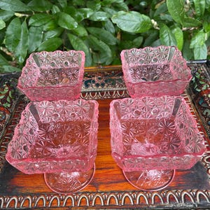 May include: Four pink glass square dishes with a decorative starburst pattern. The dishes are on a decorative wooden tray. The dishes have a scalloped edge and are elevated on a small pedestal base. The glass is a translucent pink color.