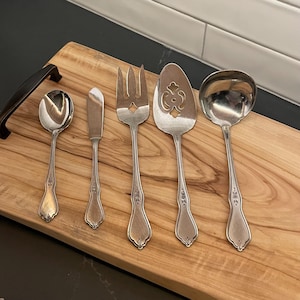 May include: Five silver serving pieces on a wooden cutting board. The pieces include a spoon, butter knife, serving fork, slotted spoon, and a serving spoon with a decorative cut-out design.
