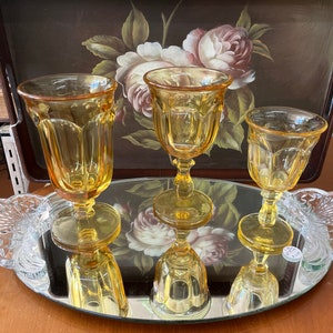 May include: Three yellow glass goblets with a fluted design sit on a mirrored tray with a floral pattern. The goblets are arranged in a triangle shape.