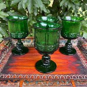 May include: Four emerald green vintage goblets with a decorative pattern on the lower half of the glass. The goblets are displayed on a wooden tray with a red and brown finish. The background features green foliage.