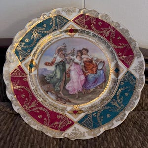 May include: A white porcelain plate with a gold rim and a colorful design. The center of the plate features a painting of three women in a landscape setting. The border of the plate is decorated with a geometric pattern in shades of red, green, and gold.