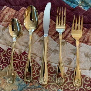 May include: Five gold-toned flatware pieces, including a knife, two spoons, and two forks, are arranged on a patterned fabric surface. The flatware features a decorative shell design on the handle.