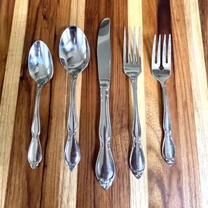 May include: A five-piece stainless steel flatware set consisting of a small spoon, a large spoon, a dinner knife, a dinner fork, and a salad fork. Each piece features a polished silver finish with a decorative floral motif at the base of the handle.