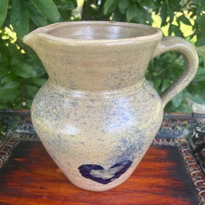 May include: A handmade, beige-colored ceramic pitcher with a spout and handle. The pitcher has a blue speckled pattern and a dark blue heart painted on the front. The pitcher is sitting on a wooden surface.