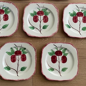 May include: A set of six square ceramic plates with a white background and a red cherry design. The plates have a scalloped edge with a red trim. The cherry design includes green leaves and a brown stem.