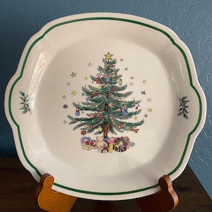 Puede incluir: Un plato navideño de cerámica blanca con borde verde y bordes festoneados. El centro presenta un árbol de Navidad decorado con adornos, regalos y un oso de peluche. Pequeñas estrellas están esparcidas alrededor del árbol.