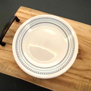May include: A round white ceramic plate featuring a decorative border pattern composed of small dark blue diamonds and a thin teal line. The plate has a smooth, glossy finish and a simple, clean design that frames the plain white center area.