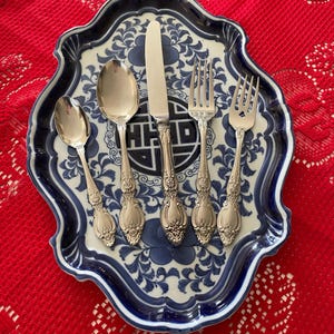 May include: A set of ornate silverware, including a spoon, knife, and forks, arranged on a blue and white patterned serving platter. The platter rests on a red, textured tablecloth. The silverware has decorative handles.