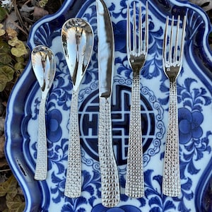 May include: A set of five silver-toned flatware pieces on a blue and white patterned plate. The set includes a knife, two forks, and two spoons.