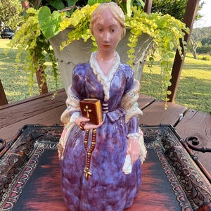 May include: A ceramic figurine of a woman in a purple dress holding a book and a rosary. The figurine is standing on a wooden tray with intricate designs.