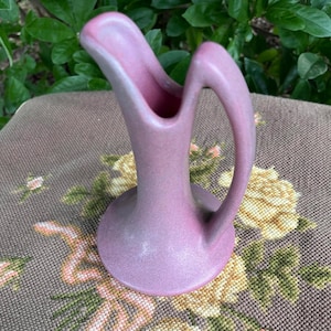 May include: A dusty rose ceramic pitcher with a smooth matte finish, featuring a wide, flared spout and a tall, curved handle that extends from the upper rim down to the base, creating a continuous, fluid silhouette for this vessel.