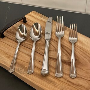 May include: A set of polished silverware, including spoons, a knife, and forks, arranged on a wooden cutting board. The silverware has a classic design with a decorative element at the base of the handles. The board has a dark handle on the left side.