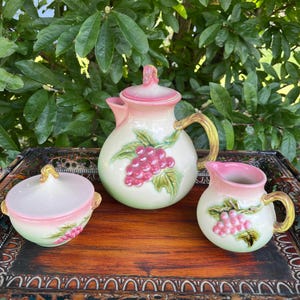 May include: A vintage ceramic tea set with a teapot, creamer, and sugar bowl. The set features a pink and white color scheme with a grape motif. The teapot has a lid and a curved handle. The creamer and sugar bowl also have handles.