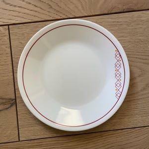 May include: A white ceramic dinner plate with a red rim and a geometric pattern in red and yellow.
