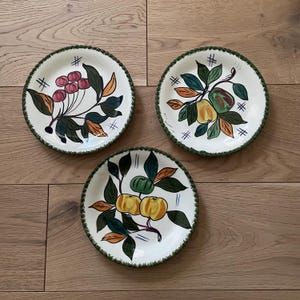May include: Three white ceramic plates with green trim and painted fruit designs. The plates feature a red cherry branch, a green apple branch, and a yellow apple branch.