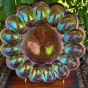 May include: A round iridescent carnival glass egg plate featuring a central circular medallion with a textured stippled pattern surrounded by twelve individual egg-shaped indentations, each displaying a vibrant purple, gold, and blue sheen.