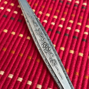 May include: Close-up of a silver fork with an ornate design of a rose and leaves. The fork is lying on a red and white patterned surface.