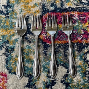 May include: Four silver forks with ornate handles, laid out on a colorful patterned rug.