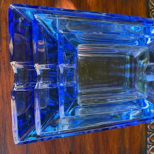 May include: A rectangular, cobalt blue glass ashtray with a stepped design. The ashtray has a square central area and a series of tiered edges. The glass is transparent, allowing light to pass through. The ashtray is placed on a wooden surface.