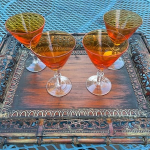 May include: Four amber-colored glass cocktail glasses with a decorative etched design sit on a wooden tray with an ornate metal trim.