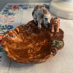May include: A brown ceramic dish with a textured surface, featuring two dog figurines and a small green frog. The dogs are white with brown spots and a smaller orange dog. The dish is on a white tablecloth with floral embroidery.