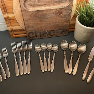 May include: A collection of stainless steel silverware, including forks, spoons, and knives, arranged on a dark surface. A wooden cheese board with the word "cheese" is in the background, along with a small potted plant.