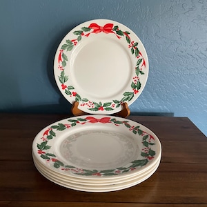 May include: Set of four white ceramic dinner plates with a green holly and red bow design. The plates are stacked on a wooden surface.
