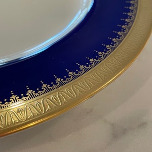 May include: Close-up of the rim of a white porcelain plate with a cobalt blue band and a gold trim. The gold trim features a repeating pattern of stylized floral and geometric designs.