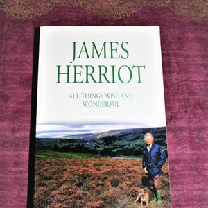 Könnte beinhalten: Ein Buchcover mit einem grünen Titel "JAMES HERRIOT" und dem Untertitel "ALL THINGS WISE AND WONDERFUL". Das Cover zeigt ein Foto eines Mannes, der mit einem Hund auf einem Feld steht.
