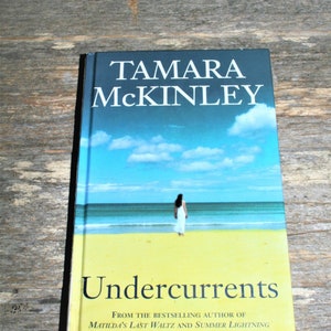 Puede incluir: Una portada de libro con un cielo azul y blanco con nubes, una mujer caminando por una playa y el título "Undercurrents" de Tamara McKinley. El subtítulo dice "De la autora superventas de Matilda's Last Waltz y Summer Lightning."
