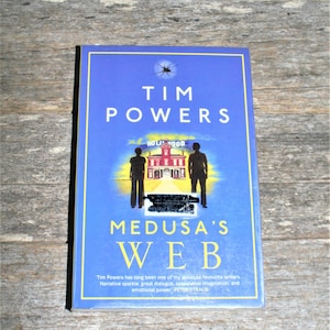 May include: A blue book cover with the title "TIM POWERS" and the book title "MEDUSA'S WEB" in yellow letters. The cover features a black and white illustration of two figures standing in front of a red building with a sign that reads "HOLLYWOOD". The text below the book title reads "Tim Powers has long been one of my absolute favourite writers. Narrative sparkle, great dialogue, speculative imagination, and emotional power - PETER STRAUB."