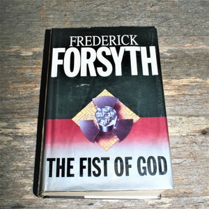 May include: A black and silver paperback book with the title "The Fist of God" by Frederick Forsyth. The cover features a red, yellow, and black design with a stylised image of a nuclear symbol.