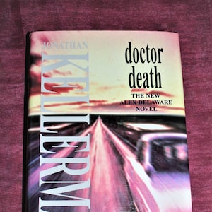 Può includere: Una copertina di libro con un'immagine sfocata di un'autostrada e il testo "doctor death the new alex delaware novel" su uno sfondo bianco. Il nome dell'autore "JONATHAN KELLERMAN" è stampato verticalmente sul lato sinistro della copertina.