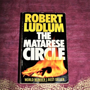 以下が含まれることがあります： 黒い表紙と黄色の文字のペーパーバック。タイトルは「The Matarese Circle」で、作者はロバート・ラドラム。表紙にはオレンジと黄色の炎の画像が描かれている。