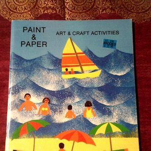 May include: A book titled "Paint & Paper: Art & Craft Activities" by David E. Corney. The cover features a sailboat on a blue ocean with waves, and people on a beach with umbrellas and beach towels.