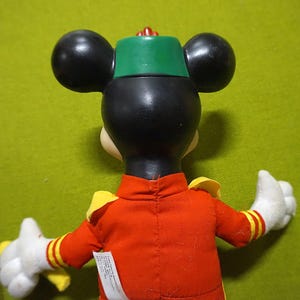 Puede incluir: Un peluche de Mickey Mouse, visto desde atr&aacute;s, con una chaqueta roja con detalles amarillos y un sombrero verde. El juguete tiene orejas negras y guantes blancos. Una peque&ntilde;a etiqueta es visible en la chaqueta.