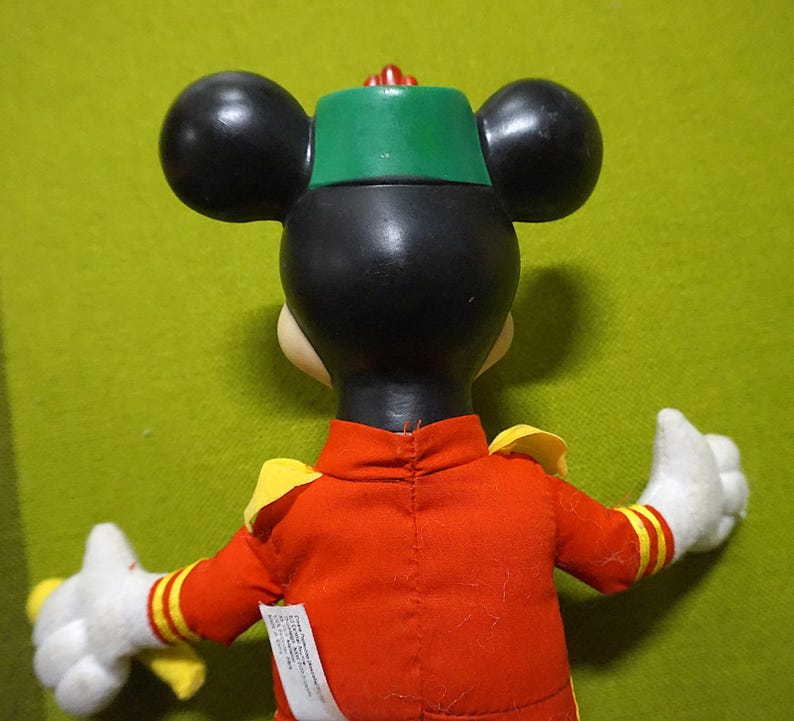 Puede incluir: Un peluche de Mickey Mouse, visto desde atr&aacute;s, con una chaqueta roja con detalles amarillos y un sombrero verde. El juguete tiene orejas negras y guantes blancos. Una peque&ntilde;a etiqueta es visible en la chaqueta.