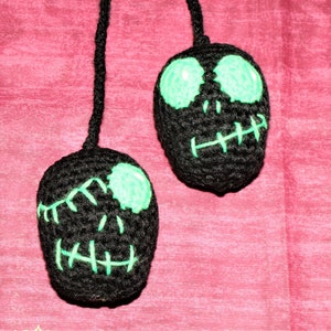 May include: Two black crocheted skull ornaments with green eyes and stitched mouths hanging from a string.