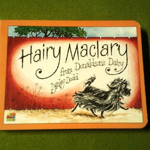 Op de afbeelding: Een kinderboekomslag met een oranje achtergrond en een zwart-witte illustratie van een hond. De titel van het boek is "Hairy Maclary from Donaldson's Dairy" door Lynley Dodd.